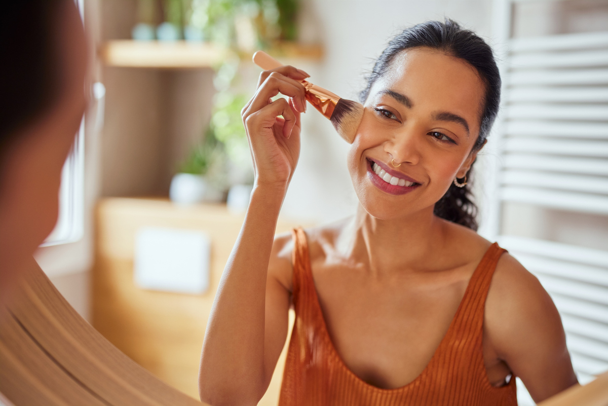 Beautiful girl applying makeup using powder brush before going to work. Healthy latin woman looking in the mirror and applying cosmetic with a big brush. Young woman looking in the mirror and applying foundation or blusher on her face.