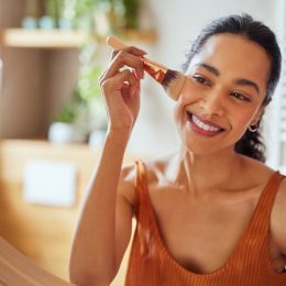 Beautiful girl applying makeup using powder brush before going to work. Healthy latin woman looking in the mirror and applying cosmetic with a big brush. Young woman looking in the mirror and applying foundation or blusher on her face.