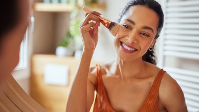 Beautiful girl applying makeup using powder brush before going to work. Healthy latin woman looking in the mirror and applying cosmetic with a big brush. Young woman looking in the mirror and applying foundation or blusher on her face.