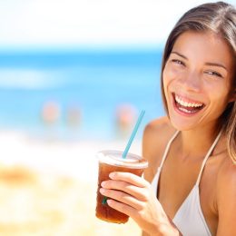 Beach woman drinking cold drink beverage having fun at beach party. Female babe in bikini enjoying Ice tea, coke or alcoholic drink smiling happy laughing looking at camera. Beautiful mixed race girl