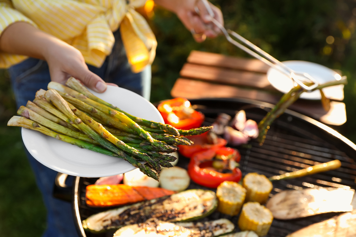Woman cooking vegetables on barbecue grill outdoors, holding out a plate of asparagus