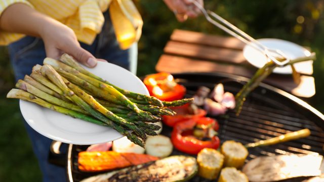 Woman cooking vegetables on barbecue grill outdoors, holding out a plate of asparagus