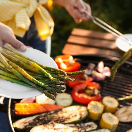 Woman cooking vegetables on barbecue grill outdoors, holding out a plate of asparagus