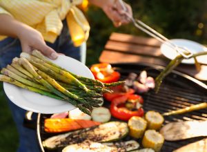Woman cooking vegetables on barbecue grill outdoors, holding out a plate of asparagus