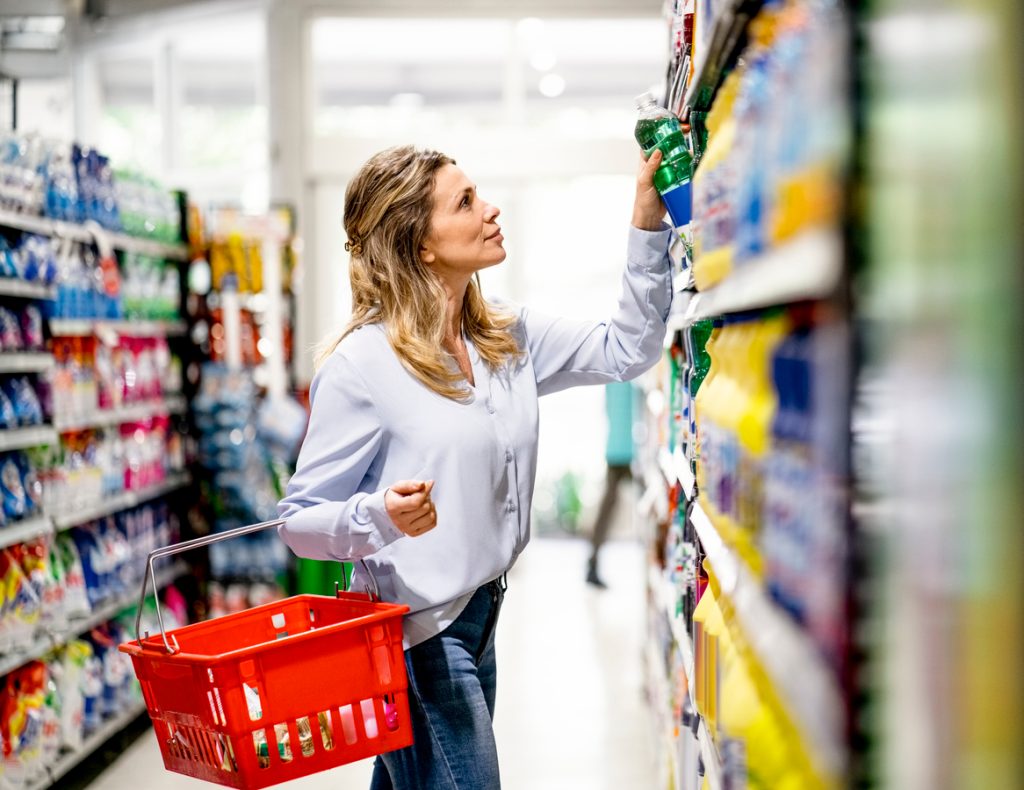 A woman grabbing a bottle from a supermarket shelf to put in her basket