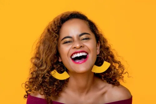 Young woman wearing a red off-the-shoulder sweater and yellow statement earrings laughing on isolated yellow background