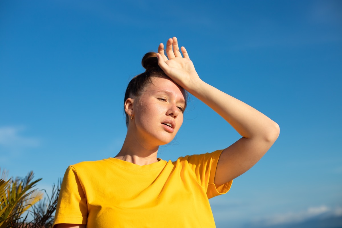 overheated young woman wearing a yellow shirt and putting a hand on her head in the sun