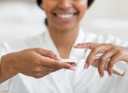 Cropped image of a smiling woman wearing a white robe putting micellar water on a cotton pad