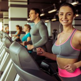 Attractive young woman in headphones running on a treadmill in gym, looking at camera and smiling