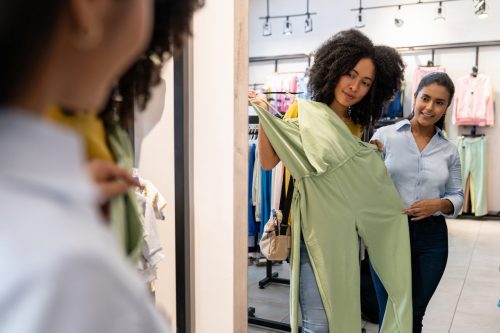Young woman holding a green jumpsuit up to a mirror while shopping