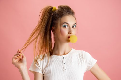 Young woman wearing a white t-shirt, blowing a bubble of yellow gum, with a side ponytail against a pink background
