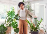 Young woman wearing orange pants and a white top waters her houseplants with a watering can