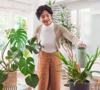 Young woman wearing orange pants and a white top waters her houseplants with a watering can