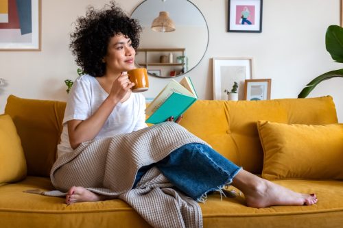 Pensive relaxed woman reading a book at home, drinking coffee sitting on the couch. Copy space. Lifestyle concept.