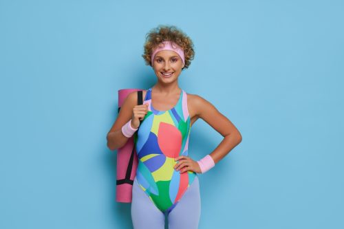 Happy woman with short haircut wearing colorful leotard and tights posing on blue background with pink yoga mat on her shoulder