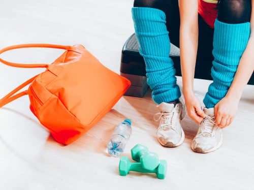 Cropped shot of woman wearing sneakers and blue leg warmers, preparing for working out at gym. Her orange gym bag is next to her, along with green hand weights and a water bottle.
