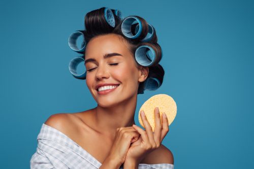 woman in a robe with large rollers in her hair holding a body sponge against a blue background