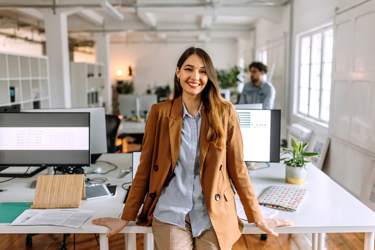 young woman in business-casual outfit leaning on a desk in an office