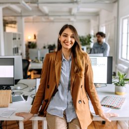 young woman in business-casual outfit leaning on a desk in an office
