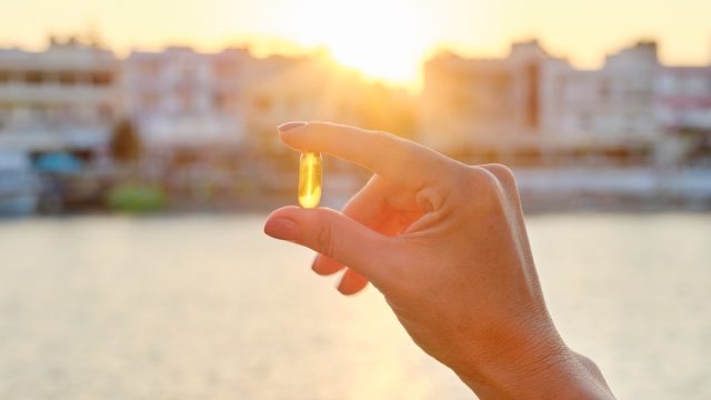 Yellow gel capsule vitamin d omega-3 in womans hand, sun sea background.