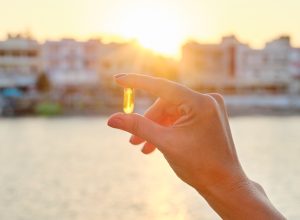 Yellow gel capsule vitamin d omega-3 in womans hand, sun sea background.