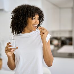 Happy woman in a white t-shirt takes a supplement with a glass of water in her kitchen