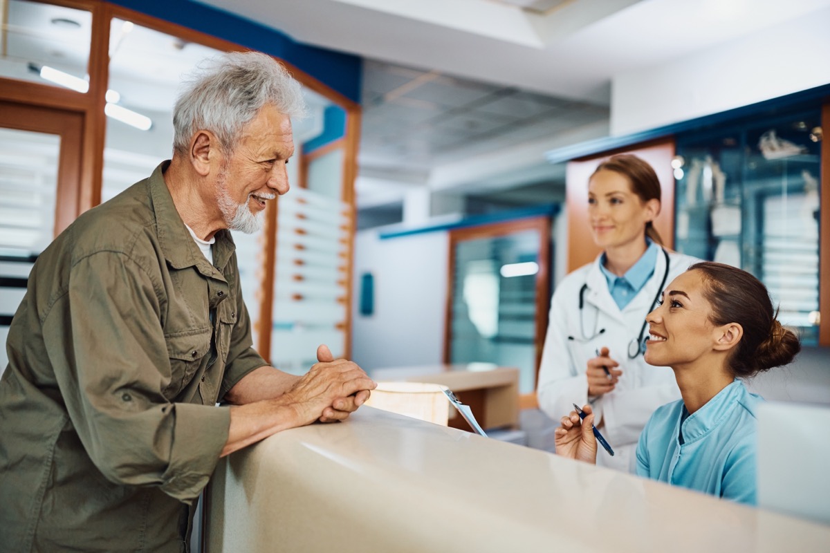 Young happy nurse taking notes while communicating with mature patient at reception desk at medical clinic.