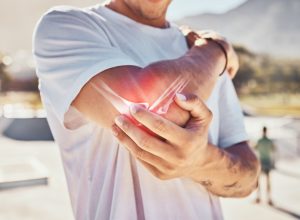 Man, joint pain and elbow arthritis in outdoor closeup in summer sunshine with massage arms. Broken arm, bone and guy at urban park in metro, city or town with physical therapy with hands in zoom