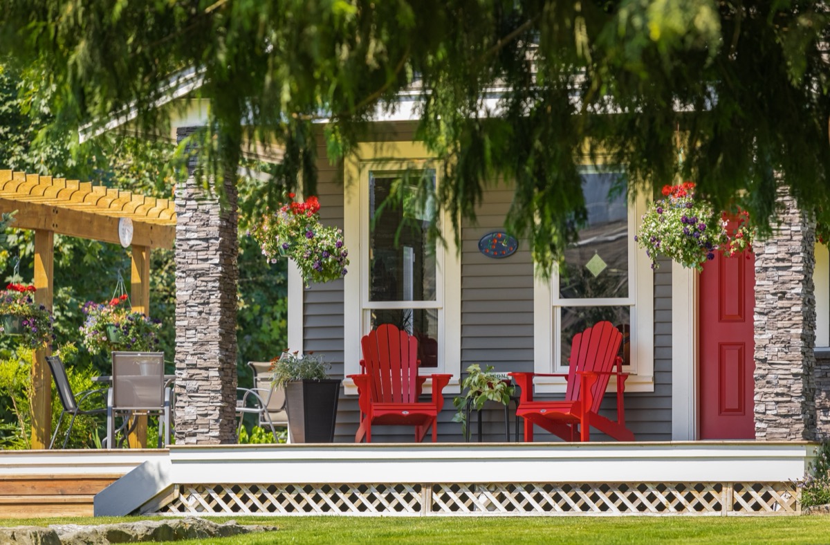 View of a large front porch with furniture and potted plants. Rustic, country style front porch seating with red Adirondack chairs and fresh flowers.