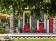 View of a large front porch with furniture and potted plants. Rustic, country style front porch seating with red Adirondack chairs and fresh flowers.