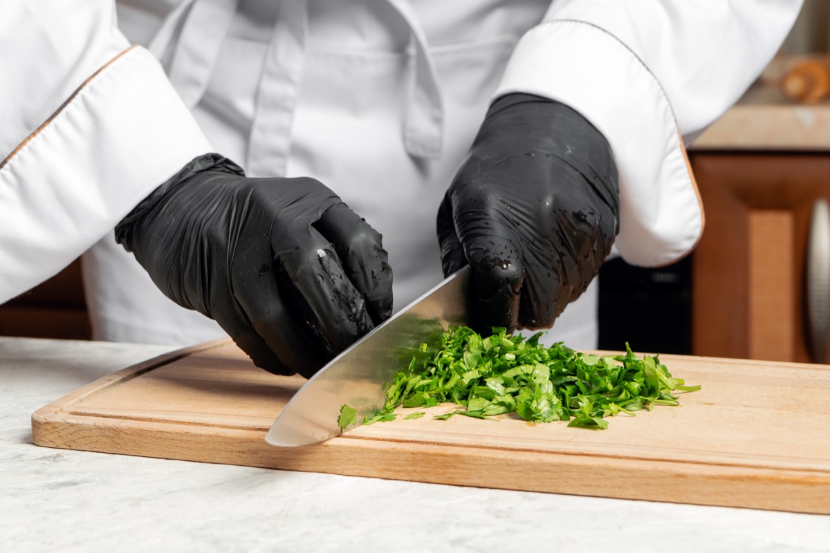 cropped image of a chef wearing a white coat and black gloves cutting parsley on a wooden board