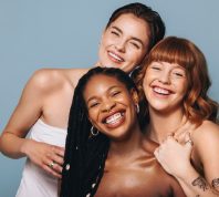 Cheerful women with different skin tones smiling at the camera in a studio. Group of happy young women embracing their natural skin. Portrait of three body positive young women standing together.