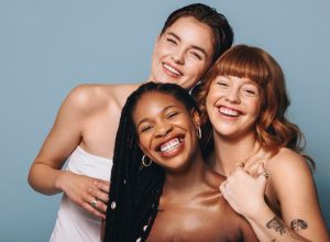 Cheerful women with different skin tones smiling at the camera in a studio. Group of happy young women embracing their natural skin. Portrait of three body positive young women standing together.