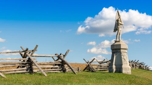A stone monument of a civil war soldier stands before a wooden split rail fence as it marks the location of Antietam National Battlefield in Sharpsburg, Maryland