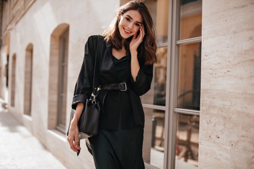Cute girl with brown wavy hairstyle, bright makeup, dark outfit of long dress, oversized jacket, belt on waist and trendy bag, standing on daylight street in city and smiling