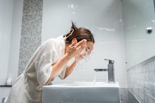 woman washing her clean face with facial foam and water. Attractive female in bathrobe washing face for healthy beauty treatments and skin care then looking at the mirror in bathroom.