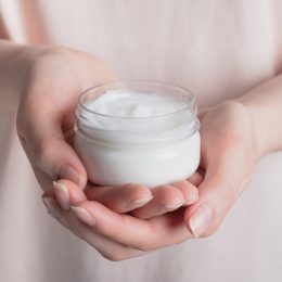 Closeup of a woman in a pale pink shirt holding out a jar of face cream