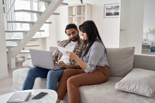 Young couple checking paper bills paying mortgage loan bank debt online together on computer, calculating taxes, expenses, making payments, planning family budget finances using laptop at home.