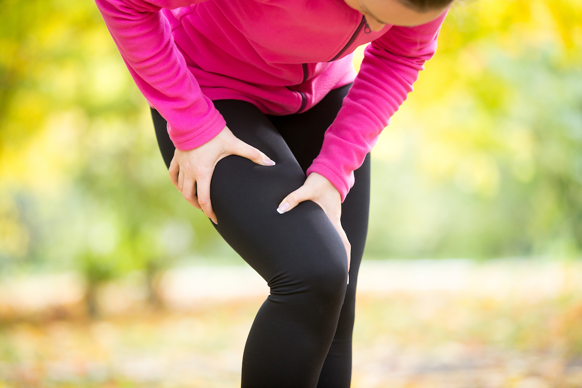 woman wearing black leggings and a pink top grabbing her leg in pain while exercising outside