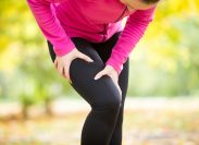 woman wearing black leggings and a pink top grabbing her leg in pain while exercising outside
