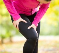 woman wearing black leggings and a pink top grabbing her leg in pain while exercising outside