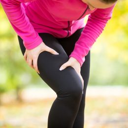 woman wearing black leggings and a pink top grabbing her leg in pain while exercising outside