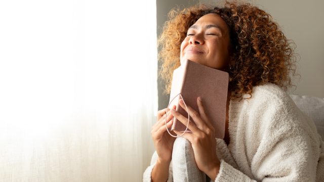 Happy woman dressed in beige smiling with her eyes closed while holding a journal