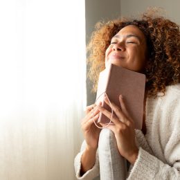 Happy woman dressed in beige smiling with her eyes closed while holding a journal