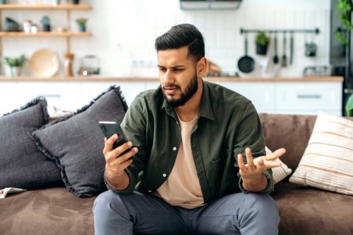 puzzled young man in casual wear, sits on sofa in living room, using mobile phone, texting online, browsing social networks, reads news, looks at screen in confusion