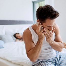 Young man sits on the edge of the bed whispering on the phone while a woman sleeps in the background, signaling cheating.