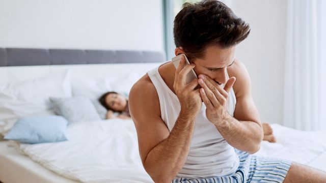 Young man sits on the edge of the bed whispering on the phone while a woman sleeps in the background, signaling cheating.