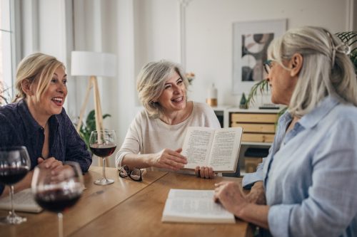 Three women sitting at a dining room table discussing a book while drinking red wine