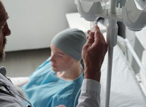 Close-up of mature male oncologist preparing dropper for chemotherapy before procedure while standing by bed with sick female patient