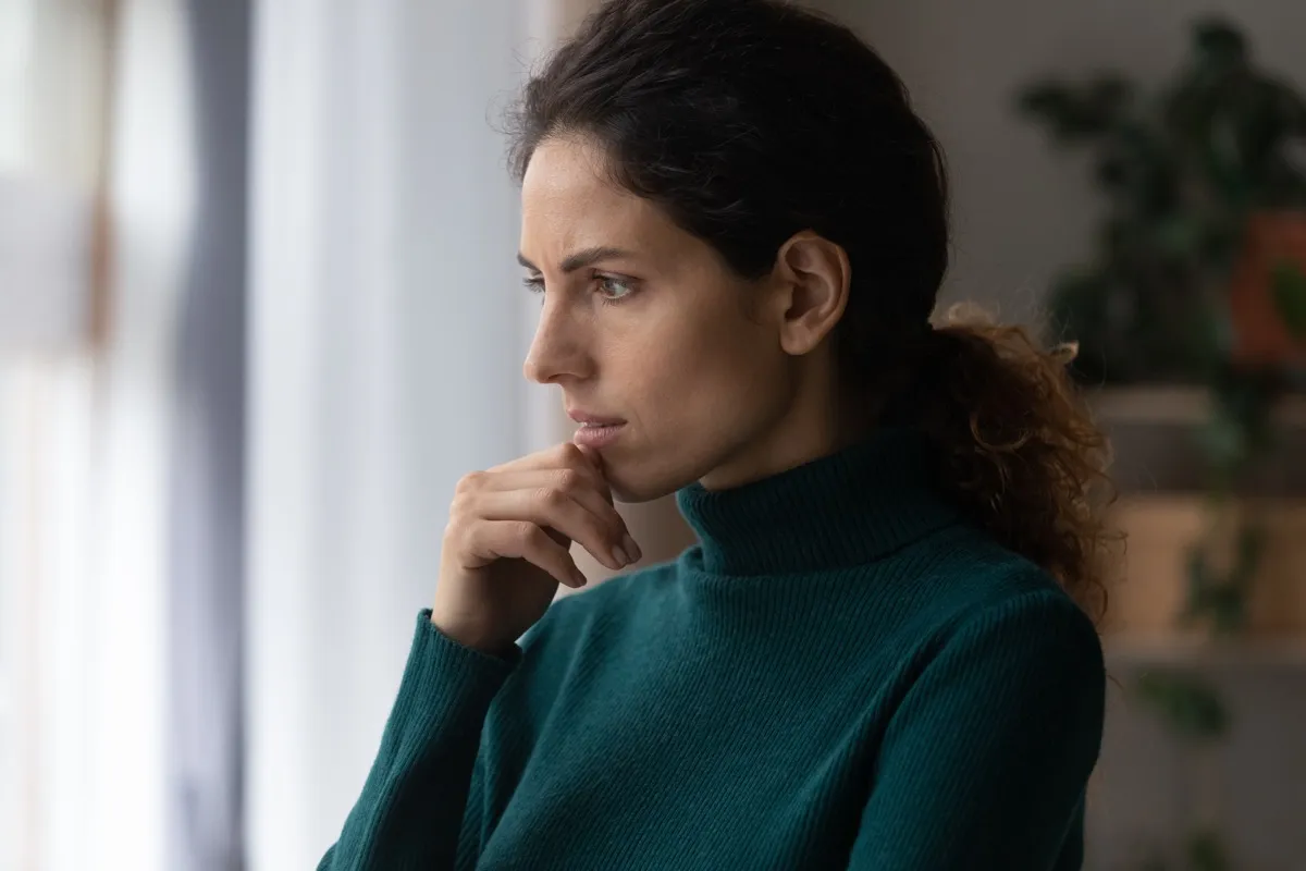 Thoughtful unhappy jealous woman touching chin, standing near window, considering problem solution or feeling stressed of difficult life situation, bad mood depression concept.